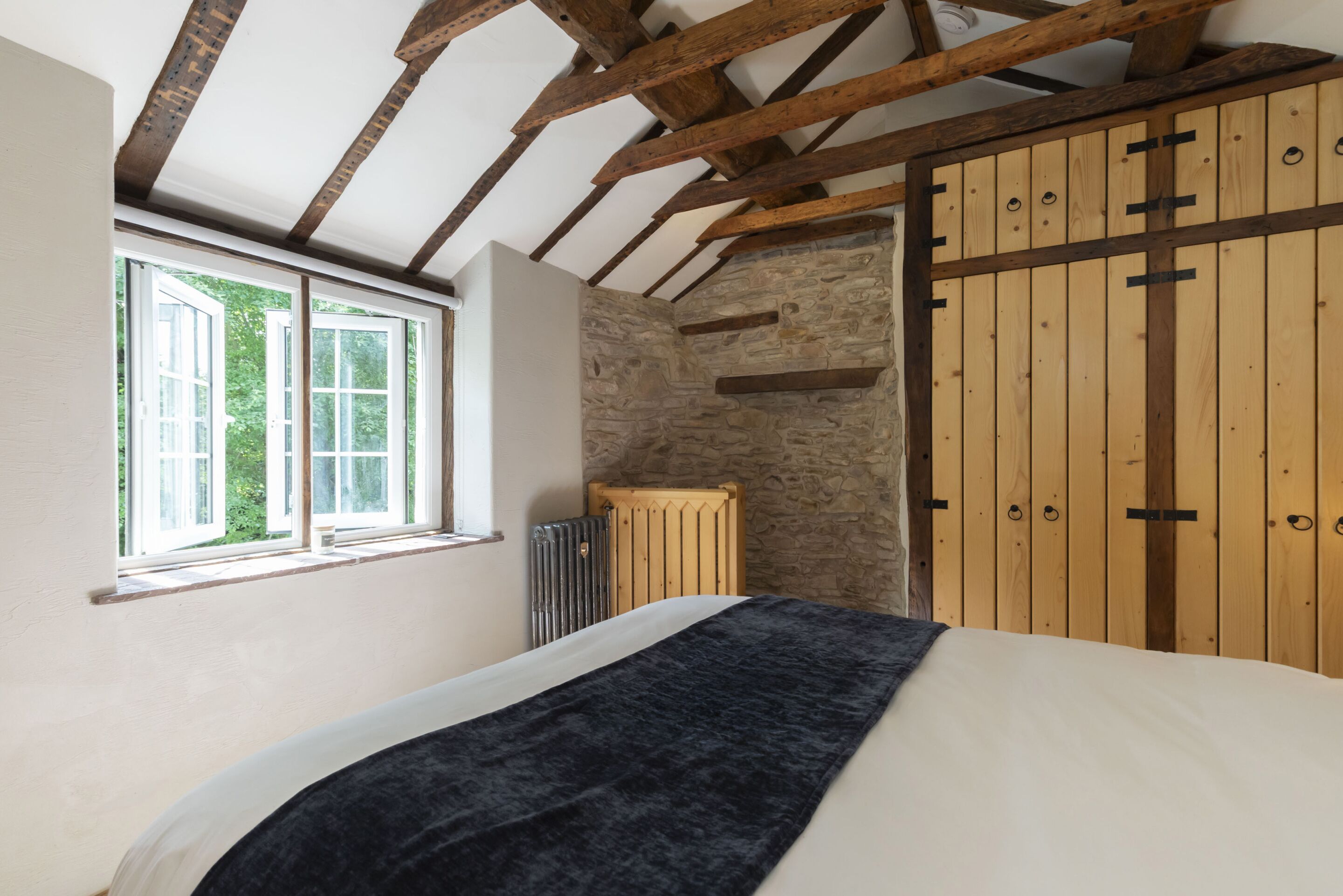 A bedroom with a large bed, wooden beams, a stone wall, and a wooden wardrobe.