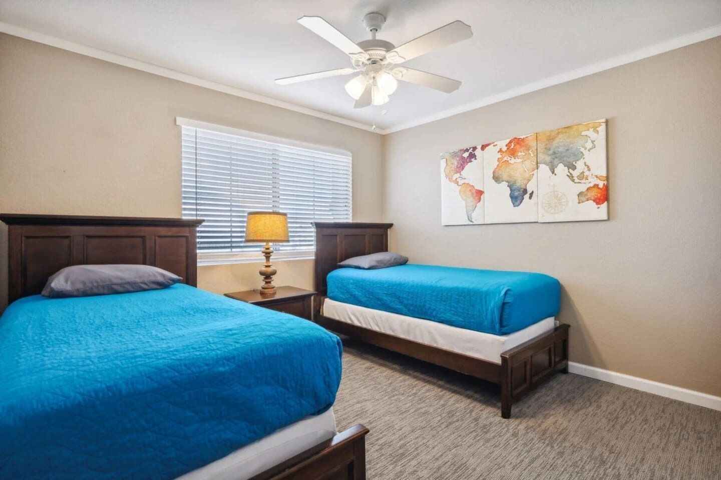 A hotel room with two beds, a ceiling fan, a window with blinds, a lamp, and a world map artwork on the wall.