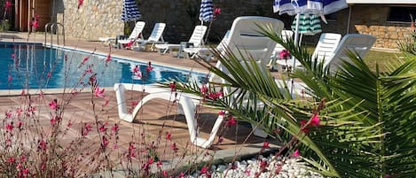 A pool area with lounge chairs and umbrellas, surrounded by a stone wall and vegetation.