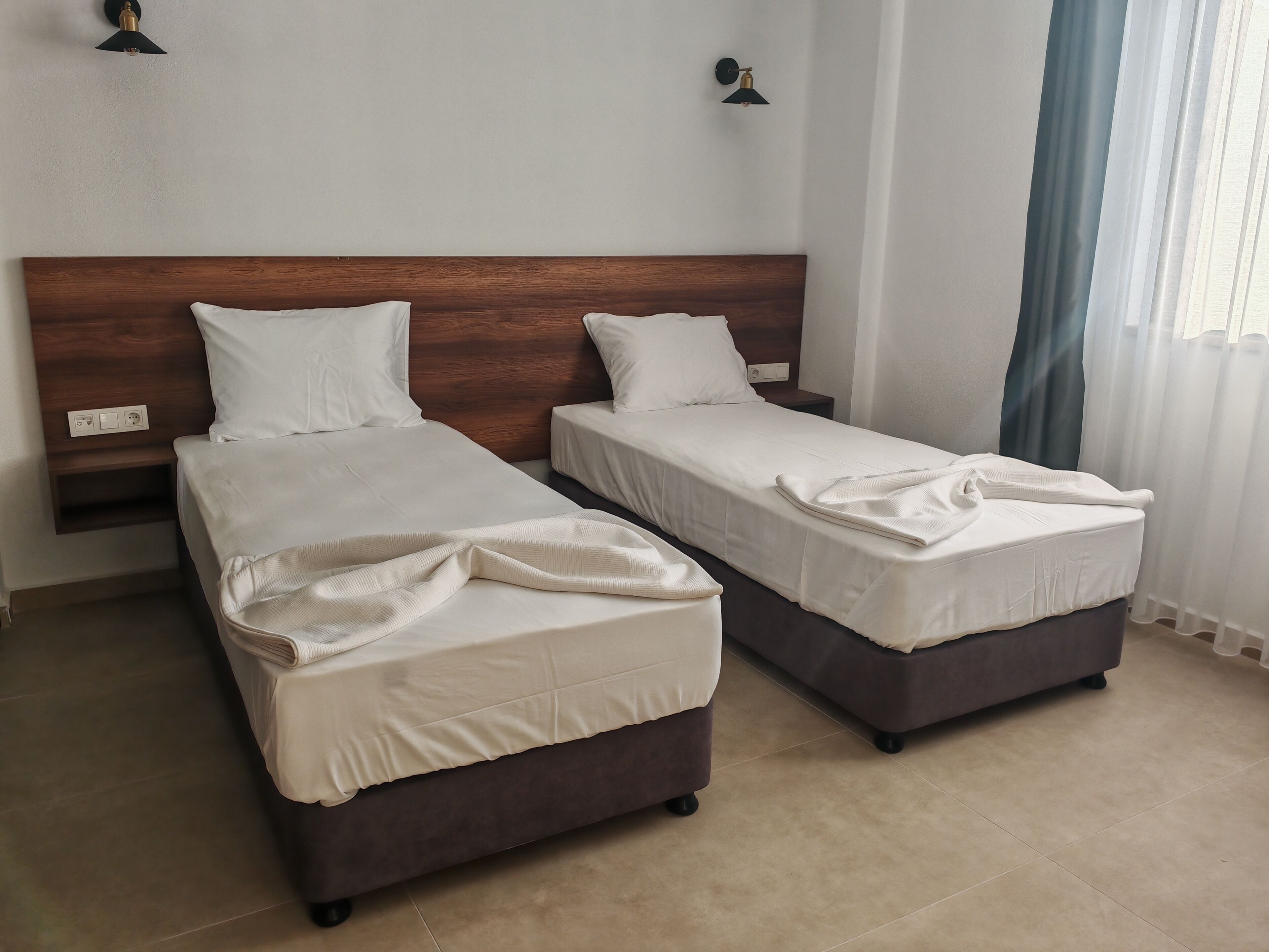 A hotel room with two single beds, a wooden headboard, a nightstand, and a window with curtains.