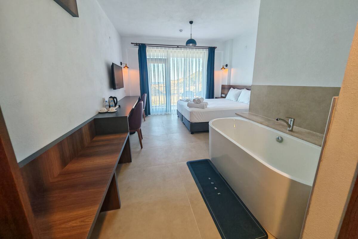 A modern hotel room with a wooden desk, a bed, a large bathtub, and a TV mounted on the wall.