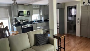 A modern kitchen with grey cabinets, a microwave, stove, and a refrigerator. There is a wooden dining table with chairs and a beige sofa in the living area. - 1.5 Blocks to Bay Beach & Pier! Huge 5BR w/ 4 Kings & 4-Car Pkg, Private Yard. (Provincetown)