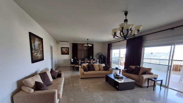 A spacious living room with a sofa set, a coffee table, and a chandelier. There is a dining area in the background with a fan and a view of the outdoors through large windows. - Belle Maison Bdadoun by Hansa  (Blaybel)
