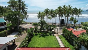 A view of a beachside property with a lawn, palm trees, and a building with a red roof. - Luxury Beachfront Retreat – 13BR Private Resort with Pool in Jacó (Jaco)