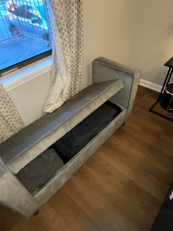 Un sofà gris amb un compartiment d'emmagatzematge integrat, col·locat en una habitació amb terres de fusta i una finestra amb cortines estampades.