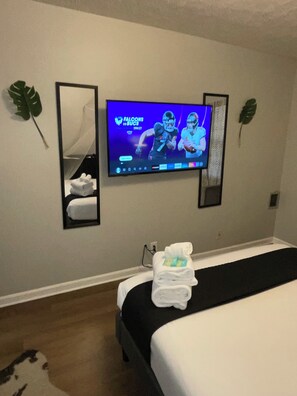 Una habitació d'hotel amb un llit, una televisió penjada a la paret, un mirall i una planta.
