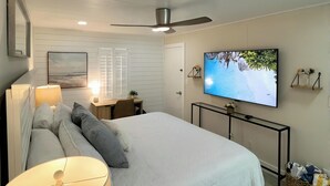 A bedroom with a bed, a ceiling fan, a TV, and a desk with a lamp.