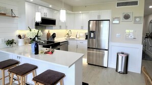 A modern kitchen with white cabinets, a stainless steel refrigerator, a large island with stools, and pendant lighting.