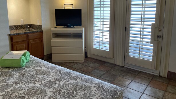 A hotel room with a kitchenette, a bed with a patterned bedspread, a television on a stand, and a sliding glass door with blinds. - Desert Paradise Resort- Gay Men's Resort (Palm Springs)