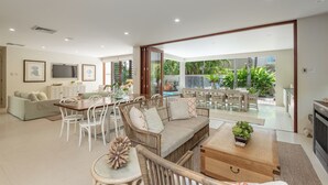 Un vaste espace de vie comprenant un canapé en rotin, une table à manger et des chaises, un téléviseur, et une vue sur le jardin et la piscine.
