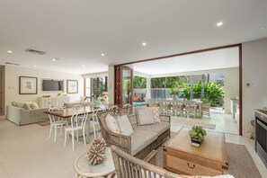 Een ruime woonkamer met een rieten bank, een eettafel met stoelen, een televisie en uitzicht op de tuin en het zwembad.