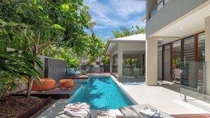 Un espace piscine extérieur moderne, doté de chaises longues, d’une petite terrasse et d’un patio fermé par des baies vitrées.