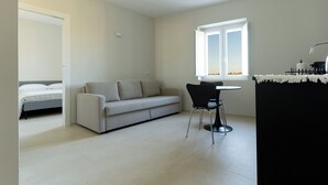 Un salon moderne et minimaliste, comprenant un canapé, une table à manger avec deux chaises, une fenêtre offrant une vue et une porte donnant sur une autre pièce.