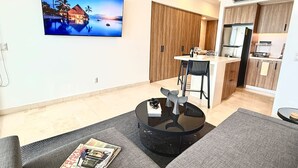 A modern living room with a flat-screen TV, a round coffee table, and a sofa. There is a kitchen area with wooden cabinets and a refrigerator.