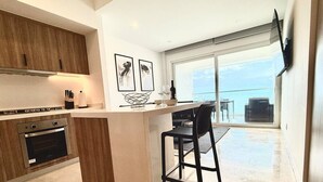 A modern kitchen with a central island, a dining area with a black chair, and a large sliding glass door leading to a balcony with a view of the sea.