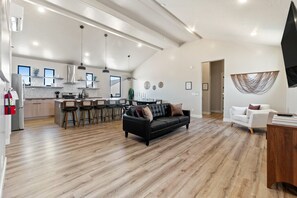 Un vaste espace de vie comprenant un canapé en cuir noir, un canapé blanc et une table à manger en bois. On aperçoit à l’arrière-plan une cuisine équipée d’appareils modernes et un coin bar.