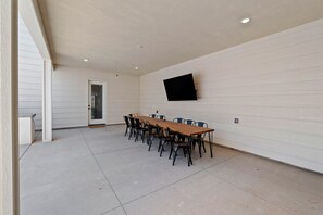Un patio cubierto con una larga mesa de madera y sillas, un televisor de pantalla plana montado en la pared y una puerta que da al interior.