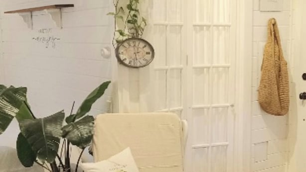 Un salon chaleureux avec un fauteuil blanc, une grande plante en pot, une horloge murale et une porte blanche.