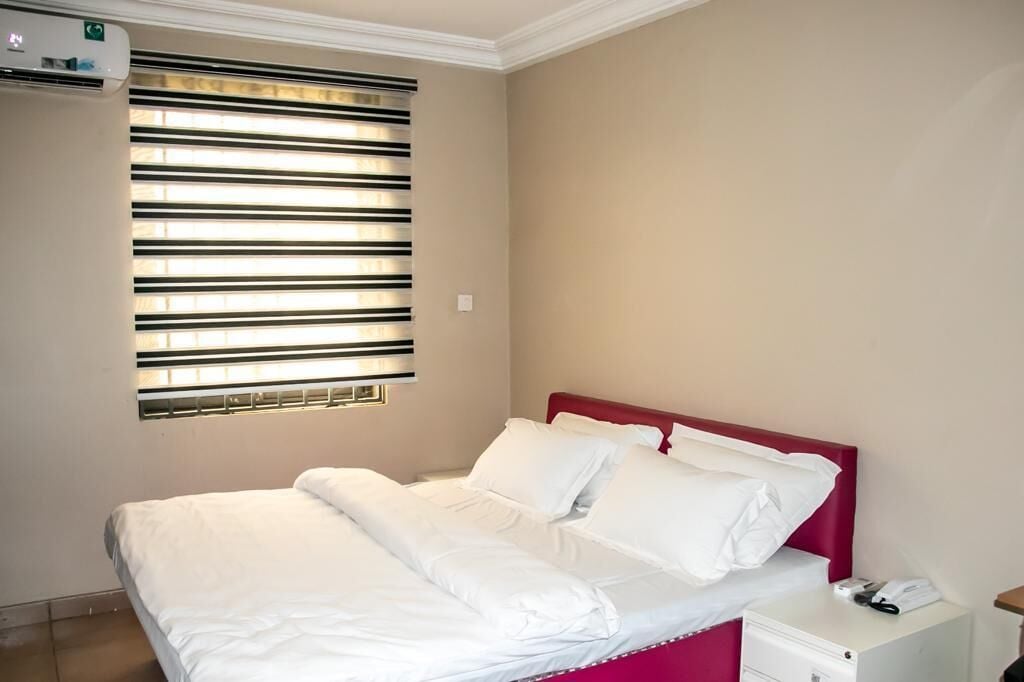 Ein Hotelzimmer mit Bett, Nachttisch, weißer Wand, Fenster mit Jalousien und Klimaanlage.