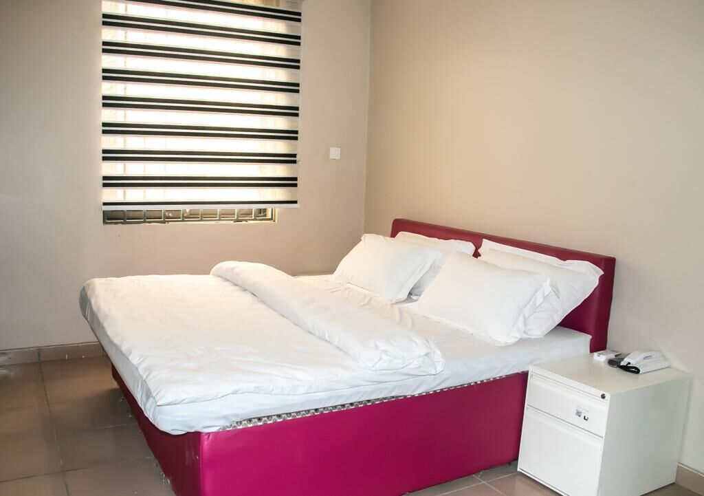 A bed with a red headboard, white bedding, and a small bedside table with a phone and a book.