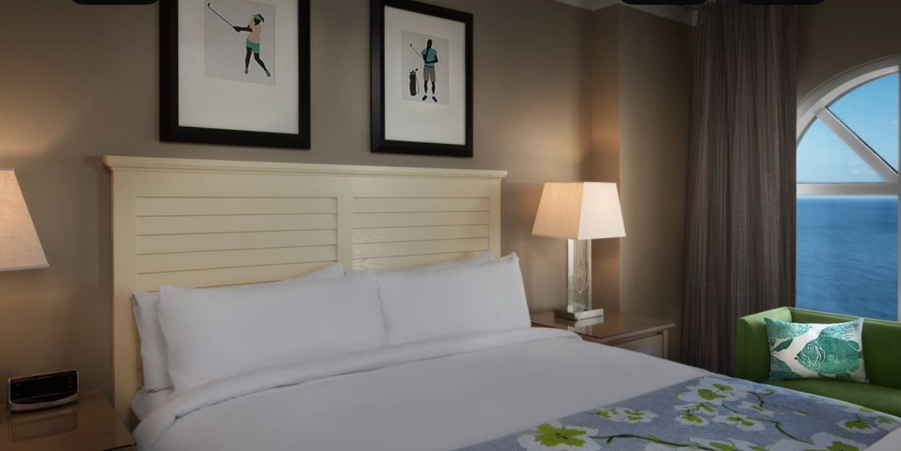 A hotel room with a large bed, two framed pictures on the wall, a green armchair, and a view of the sea.