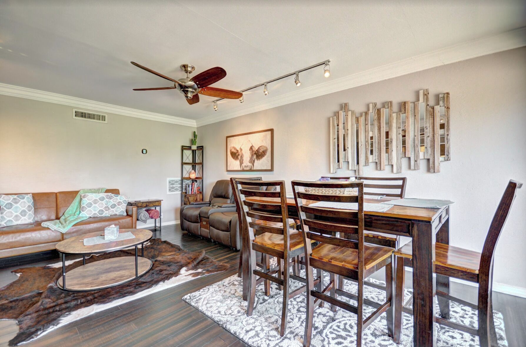 Un salon avec un canapé en cuir marron, une table basse, une table à manger avec des chaises, un ventilateur de plafond et un mur recouvert de lambris en bois verticaux.
