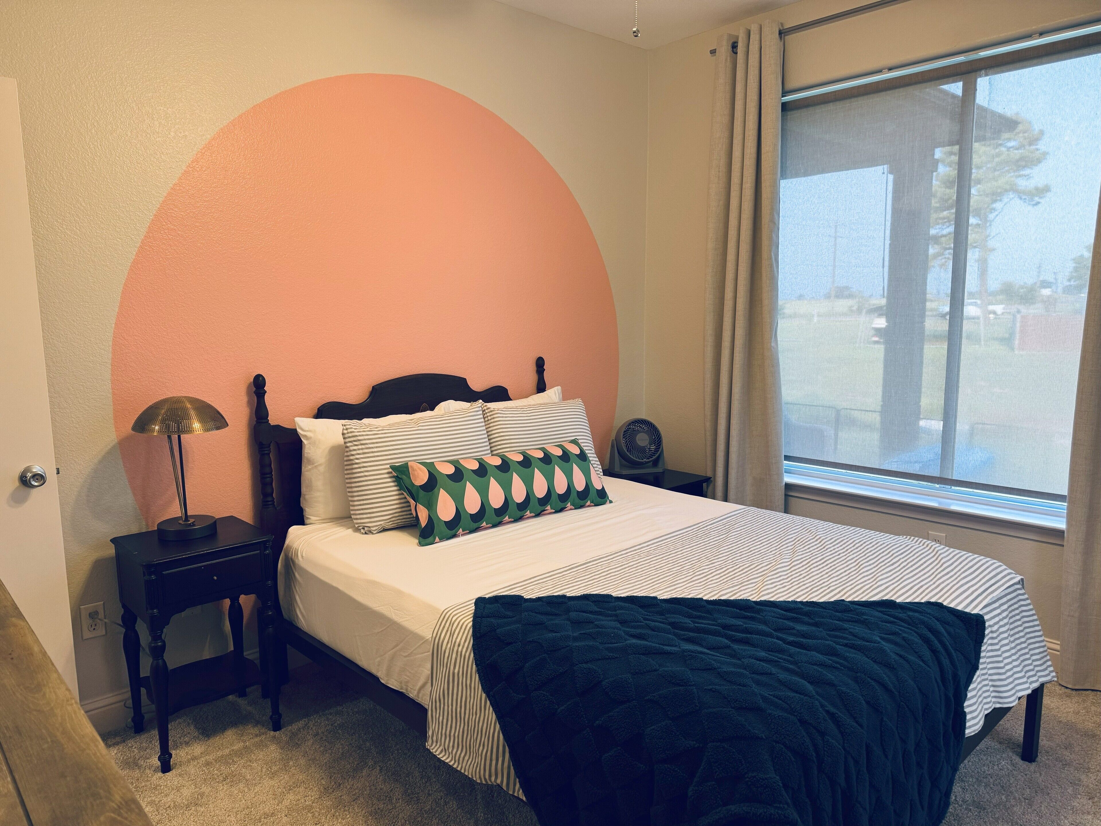 Un dormitorio con cama, mesita de noche, lámpara, ventilador y ventana con vista al exterior.