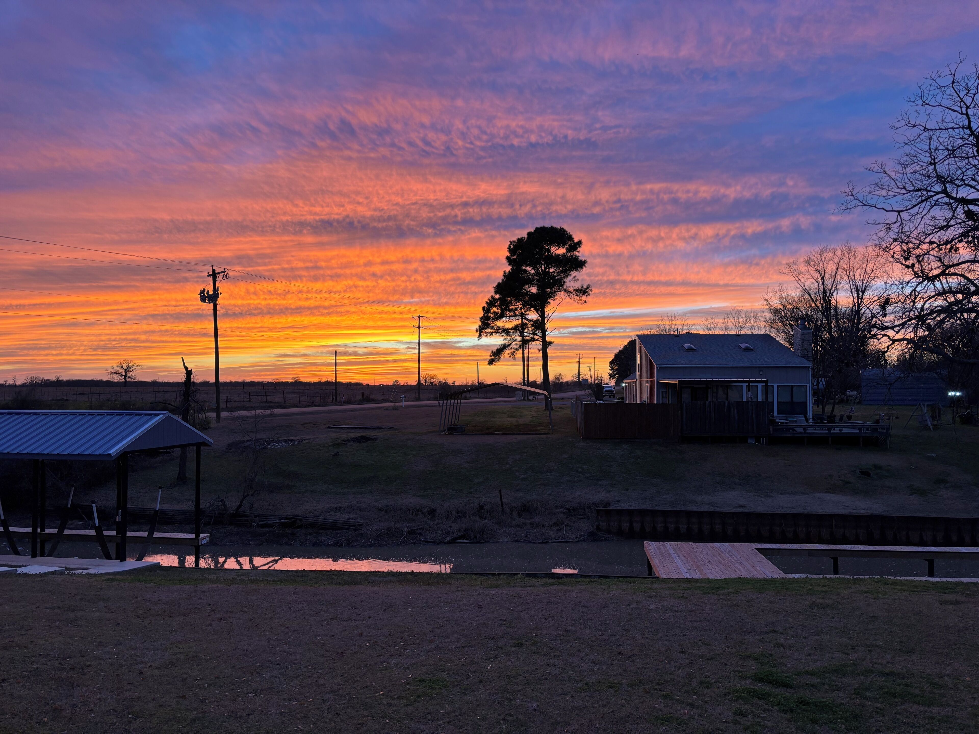Een landelijk landschap met een huis, een houten constructie en een omheind gebied tijdens zonsondergang.