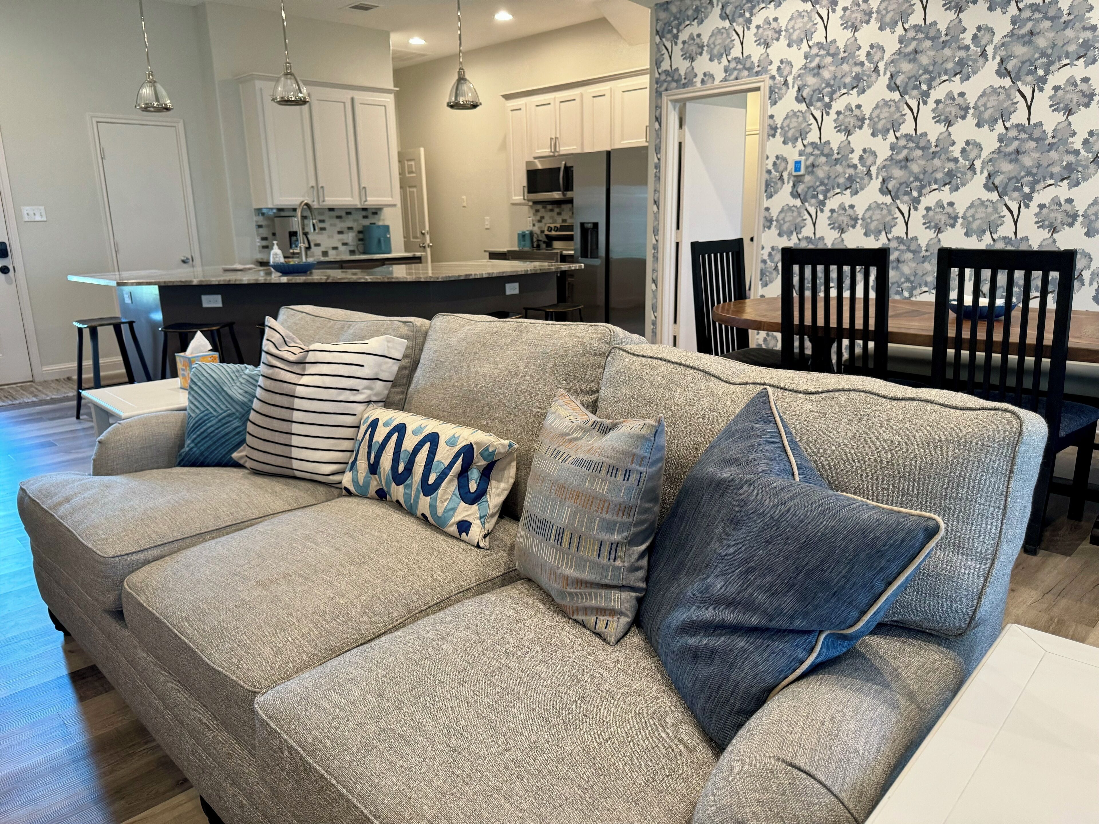 A modern living room with a beige sectional sofa, a dining area with a wooden table and chairs, and a kitchen in the background.