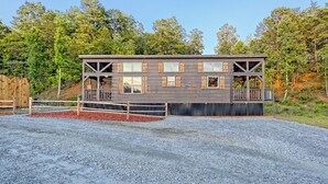 A modern wooden cabin with multiple windows and a balcony, surrounded by a gravel driveway and a wooden fence.