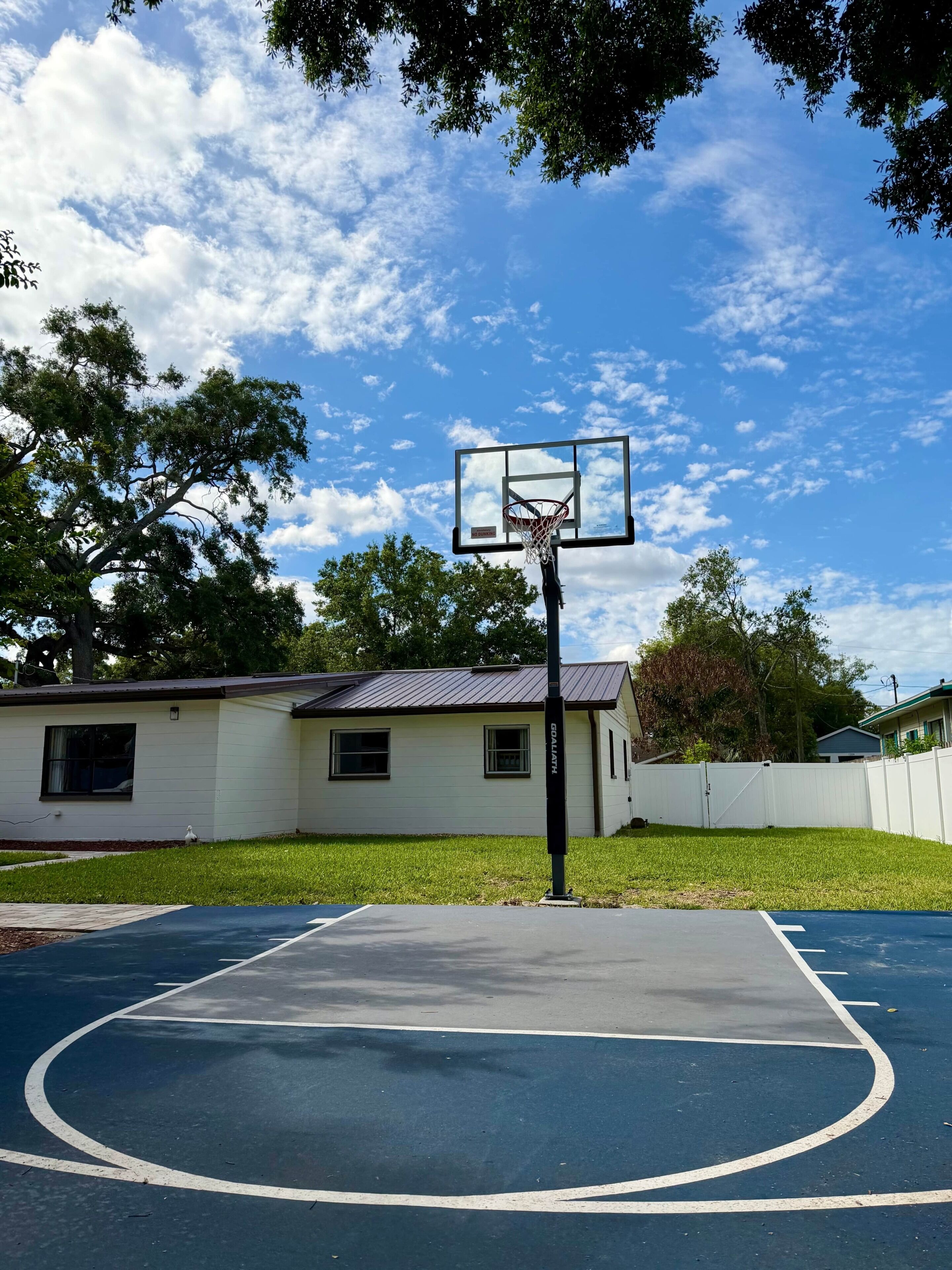 An outdoor basketball court with a hoop, surrounded by a grassy area and trees.