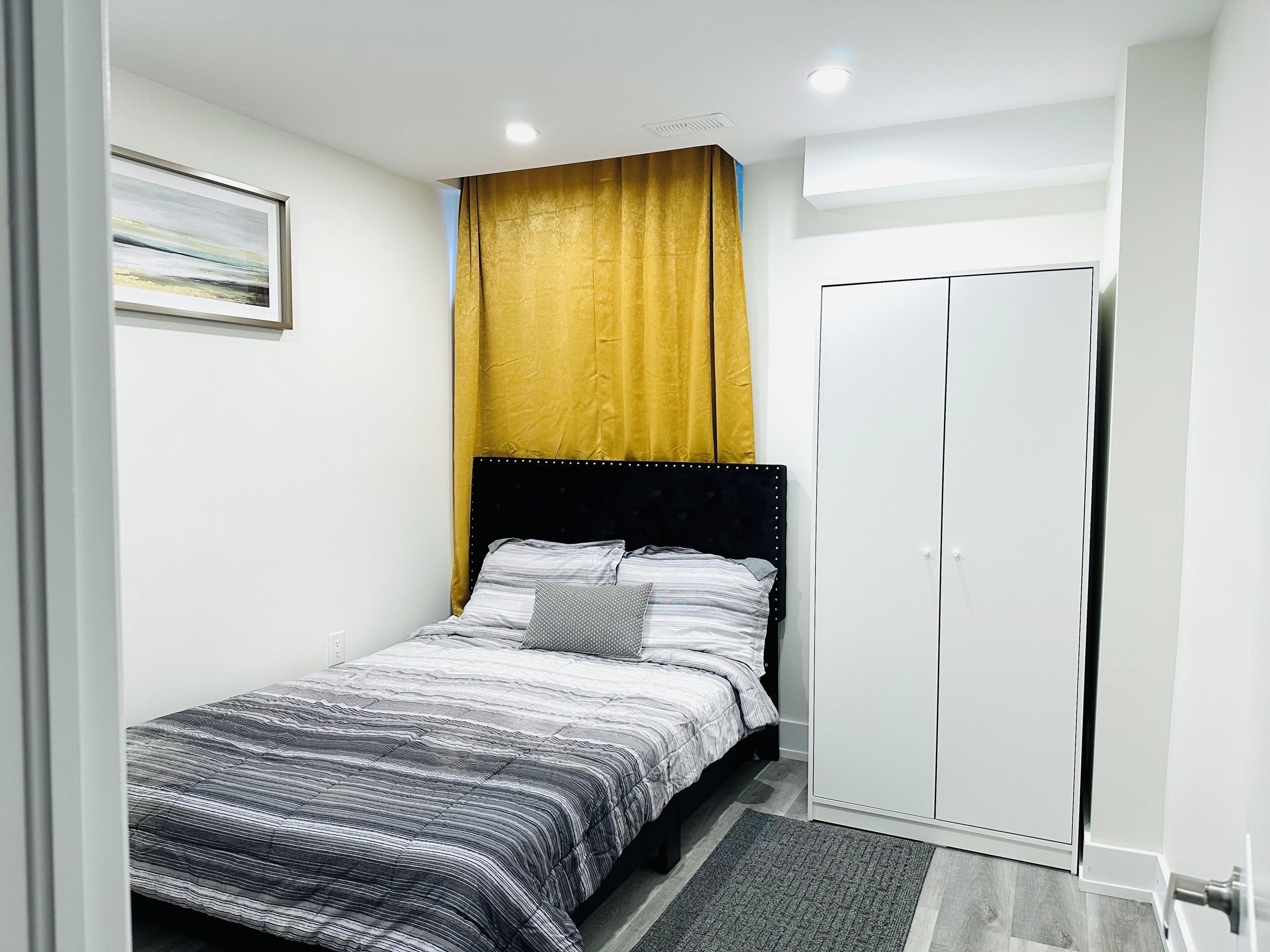 A bedroom with a bed, a wardrobe, a painting on the wall, and a ceiling light.