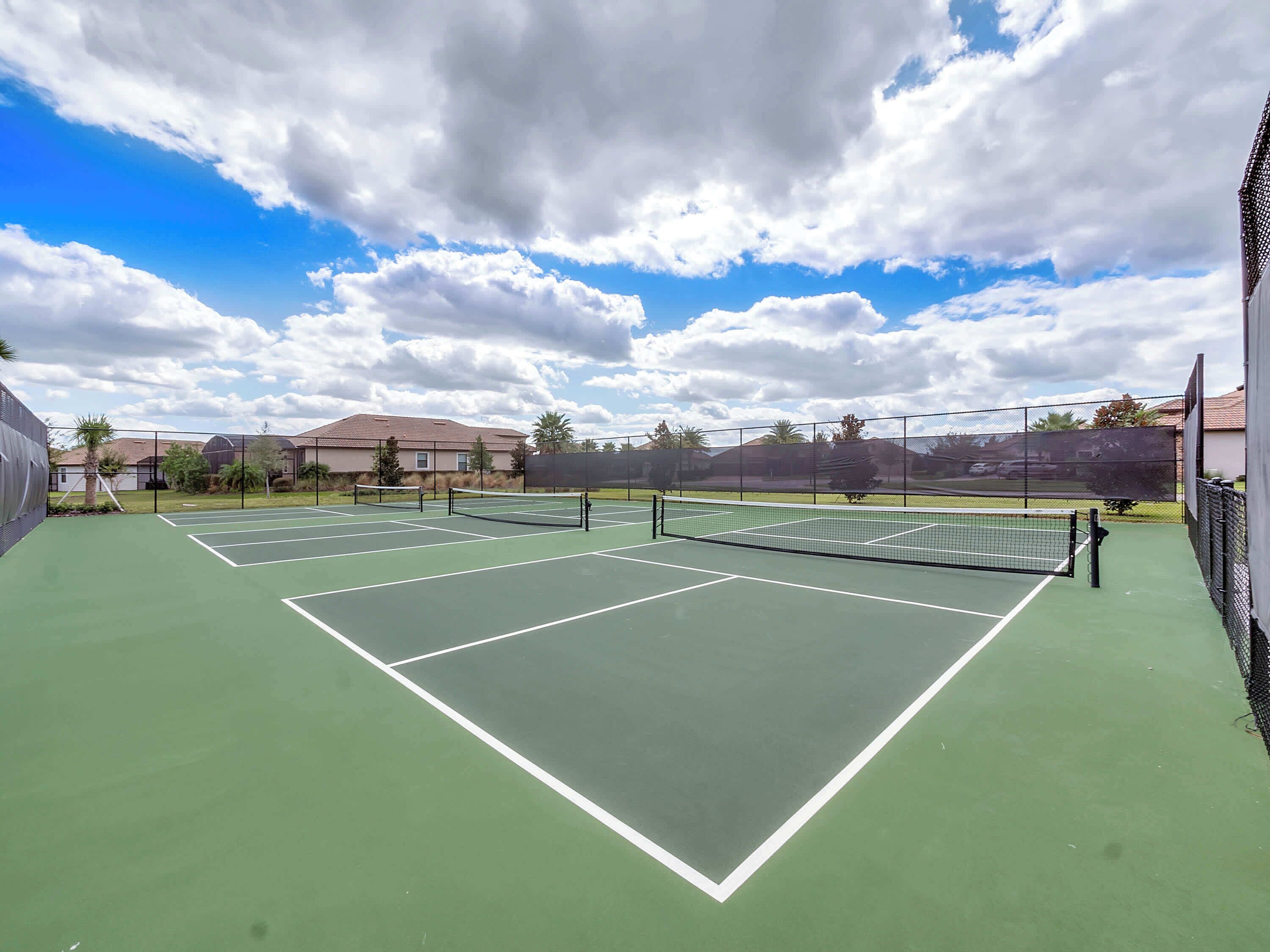 Un court de tennis comprenant plusieurs terrains, entouré d’une clôture, avec des maisons en arrière-plan.