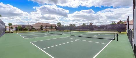 Un court de tennis comprenant plusieurs terrains, entouré d’une clôture, avec des maisons en arrière-plan.