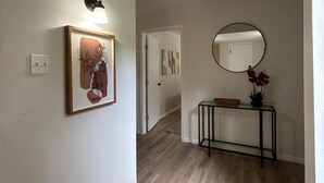 Un couloir avec un interrupteur, un tableau encadré, une console avec un miroir et une plante en pot.