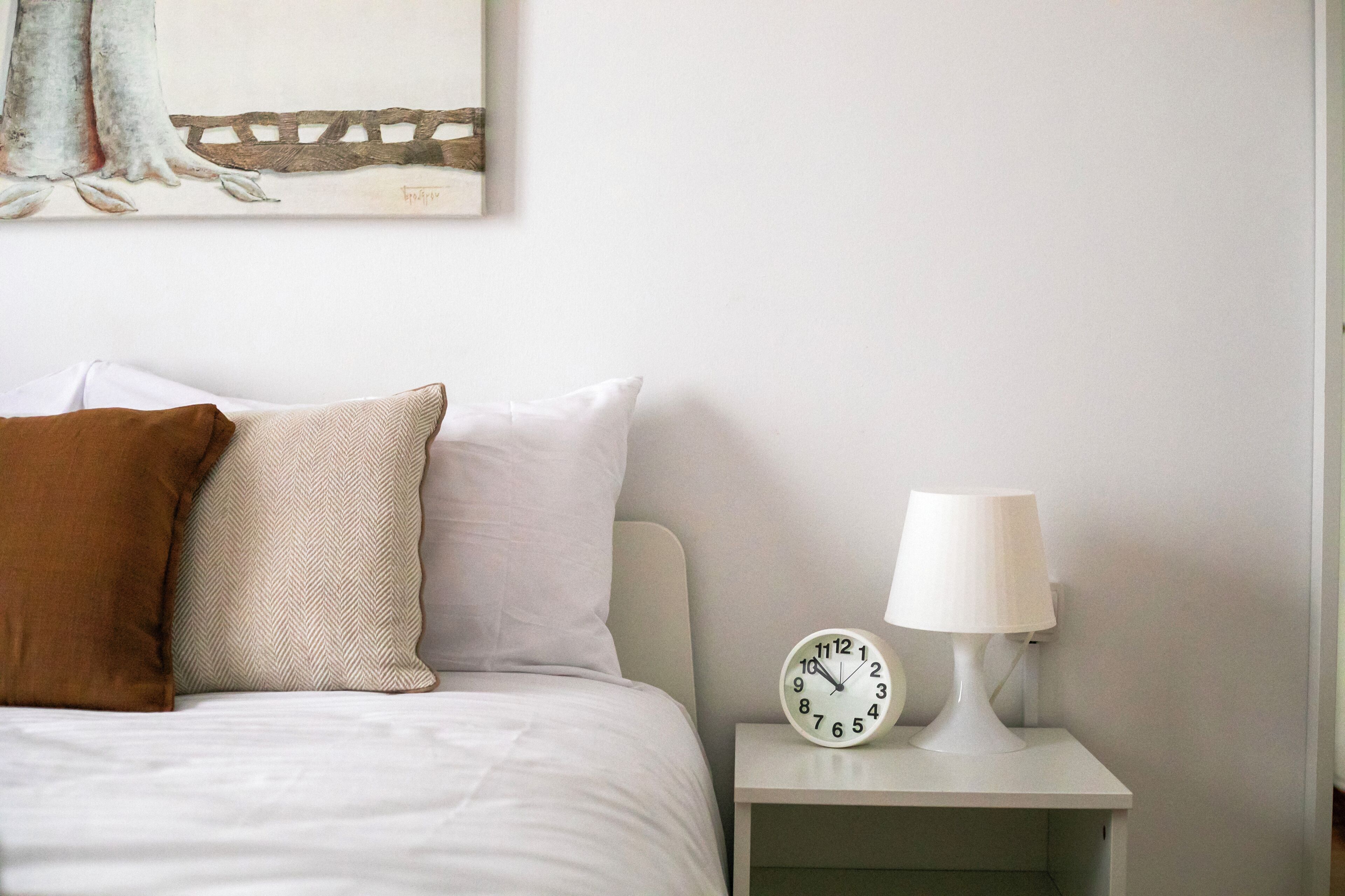 Una cama bien hecha con un almohadón blanco y marrón, una mesita de noche blanca con una lámpara blanca, y un reloj de pared.