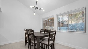 Un coin repas avec une table et des chaises en bois foncé, un lustre moderne et de grandes fenêtres équipées de stores.