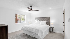 Une chambre à coucher comprenant un lit, un ventilateur de plafond, une fenêtre avec des stores et une table de chevet avec une lampe.