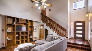 A spacious living room with a large gray sectional sofa, a wooden bookshelf, a flat-screen TV, and a wooden staircase. - Cozy Home w/Private Patio, KingBed, Fireplace, BBQ (El Paso)