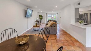 Uma sala de estar moderna com um sofá, uma mesa de jantar com cadeiras, uma TV na parede e uma área de cozinha com geladeira.