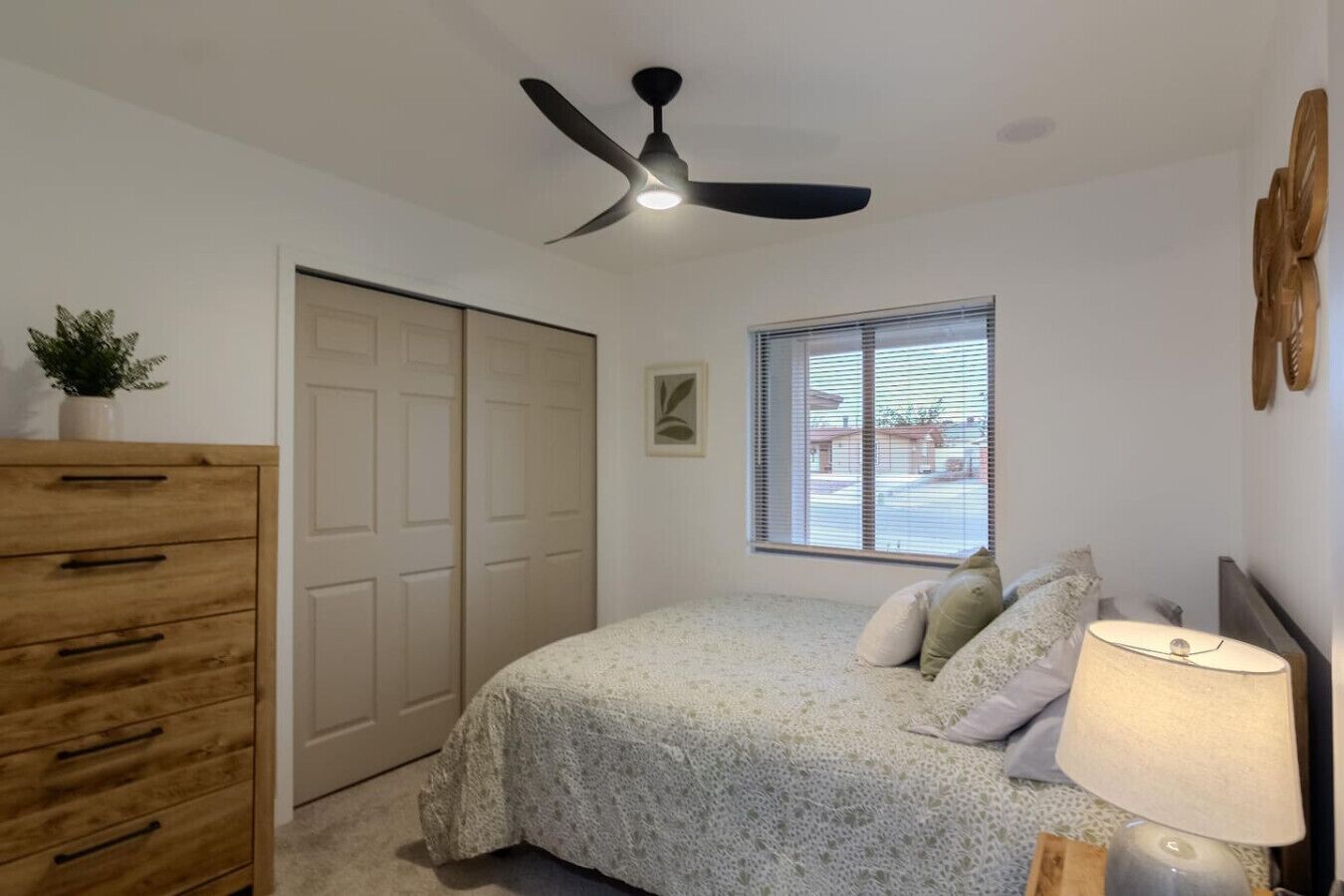 Un dormitorio con una cama, ventilador de techo, ventana con persianas, cómoda y lámpara.