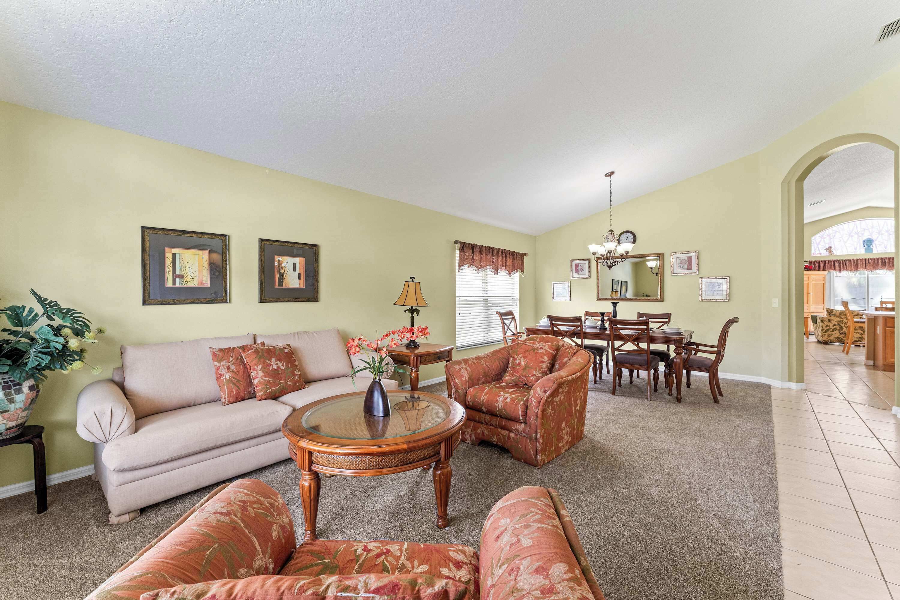 A living room with a beige sofa, floral armchairs, a coffee table, and a dining area with a table and chairs.