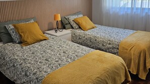 A hotel room with two beds, a bedside table with a lamp, and a large window with sheer curtains. - 7km from the beach and 14km from downtown Lisbon. Cozy, family-friendly apartment! (Almada)