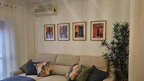 A living room with a beige sofa, patterned cushions, and a blue throw blanket. There are three pendant lights hanging from the ceiling, and a potted plant on the right. - 7km from the beach and 14km from downtown Lisbon. Cozy, family-friendly apartment! (Almada)