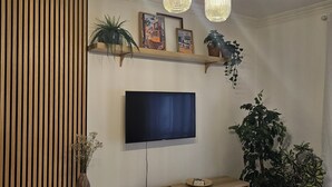 A modern living room with a flat-screen TV mounted on the wall, wooden slatted wall paneling, a white entertainment unit, and hanging pendant lights. - 7km from the beach and 14km from downtown Lisbon. Cozy, family-friendly apartment! (Almada)