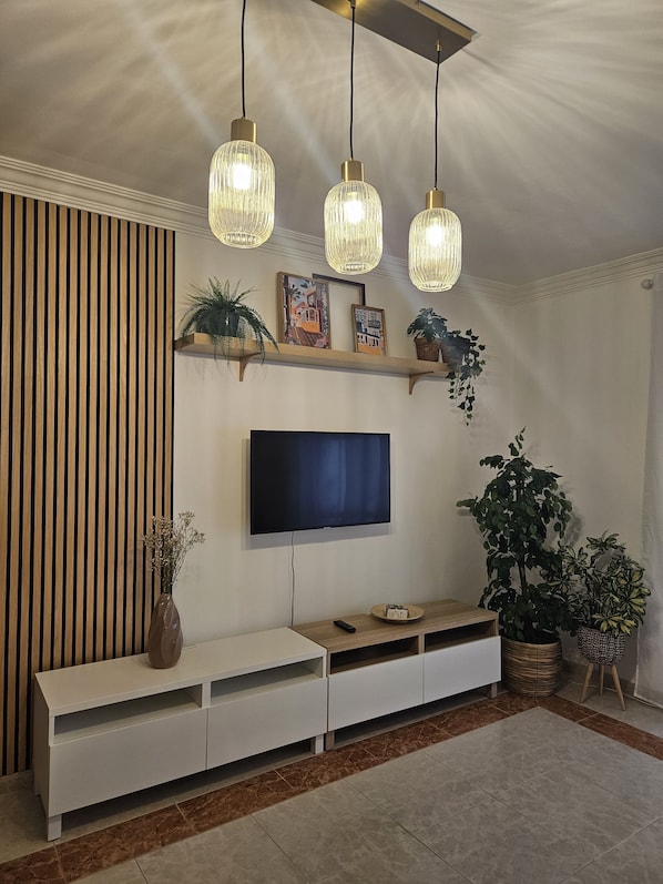 A modern living room with a flat-screen TV mounted on the wall, wooden slatted wall paneling, a white entertainment unit, and hanging pendant lights.