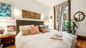 A bedroom with a large bed, multiple pillows, a nightstand, a lamp, a framed painting, a mirror, and a window with curtains.