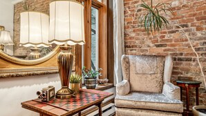 A cozy room with a vintage armchair, a wooden table with a chessboard, and a large mirror reflecting a brick wall.