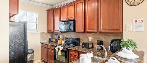 A kitchen with wooden cabinets, granite countertops, a black refrigerator, a microwave, a stove, and a sink. There is a clock on the wall and a 911 emergency contact sign.