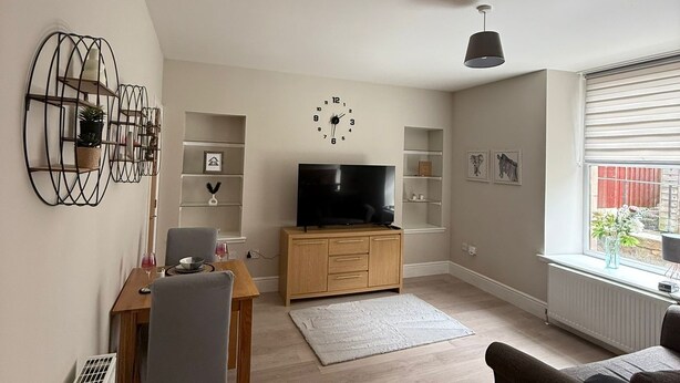 A modern living room with a flat-screen TV on a wooden cabinet, a dining table with chairs, a sofa, and a large window with blinds.